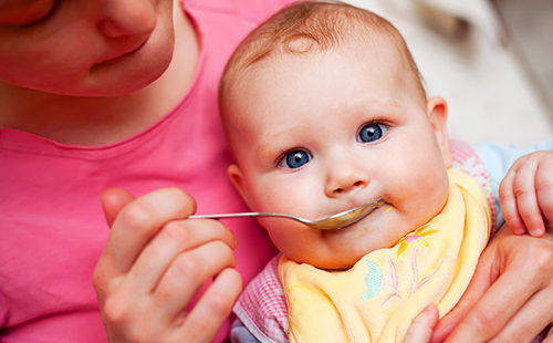 Младенец в жёлтом слюнявчике хитро смотрит и ест кашу с ложечки