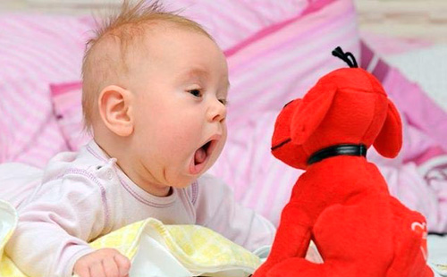 Младенец с открытым ртом смотрит на игрушечную собаку красного цвета