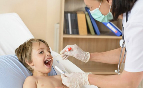 Медсестра в маске смотрит горло больному ребёнку