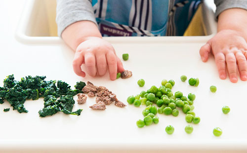 Малыш берет руками кусочки мяса и зеленый горошек