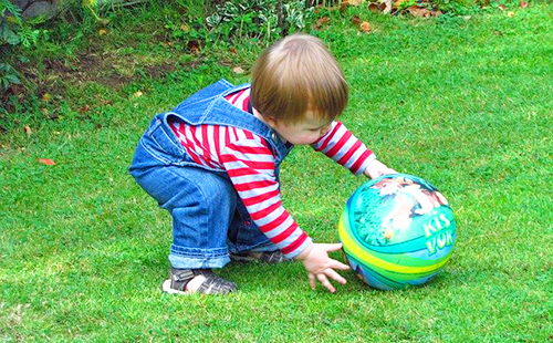 Малыш в джинсовом комбинезончике играет на лужайке с мячом